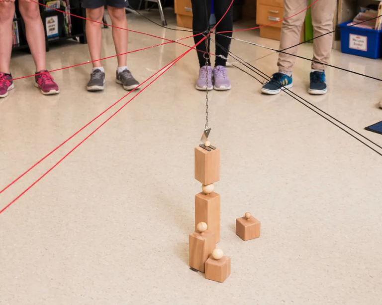 Schulkinder steuern im Klassenzimmer gemeinsam ein Kugeltower-Kooperationsspiel mit Seilen und Holzbausteinen