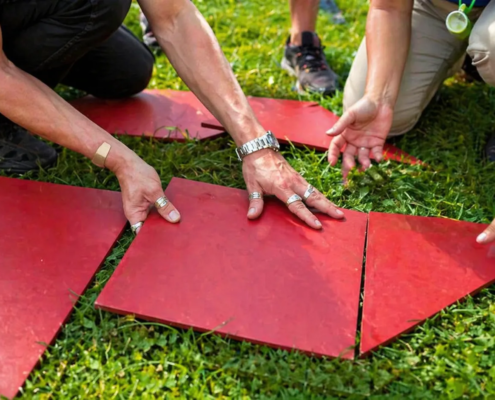 Teambuilding mit Holzspielen, Mehrere Personen legen große rote Tangram-Teile gemeinsam auf einer Wiese zusammen